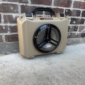 Puede incluir: Altavoz portátil de color beige con rejilla y asa negras. El altavoz tiene una superficie texturizada y la palabra "VAULT" está impresa en la parte superior. El altavoz está sobre una superficie de hormigón frente a una pared de ladrillos.