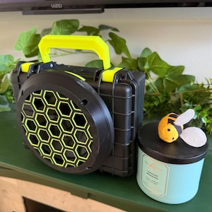 May include: Black and yellow portable speaker with a honeycomb speaker cover and handle. A teal candle with a black lid and a felt bee decoration are next to the speaker. The candle has the text "Lakeside Morning".