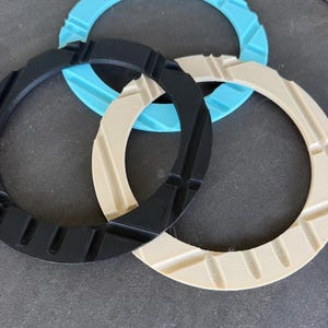 May include: Three circular rings in black, light blue, and cream colors. Each ring features a series of evenly spaced, recessed vertical grooves along its outer edge, creating a textured, gear-like appearance with a flat, smooth inner surface.