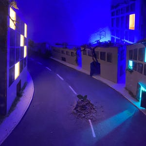May include: A miniature city scene with a road and buildings, illuminated by warm and cool-toned lights. The buildings have windows that glow, and the road has a white stripe down the center. A small plant grows in the middle of the road.