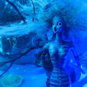 May include: A blue-toned, skeletal mermaid doll with flowing blue hair and an open-mouthed expression. The doll has a detailed ribcage design and is set against a blue, aquatic-themed background with a yellow citrus juicer.