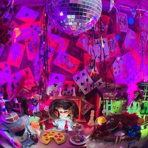 May include: A whimsical diorama scene with playing cards, dolls, and a disco ball under purple and pink lighting. A pizza and other food items are on a table. A robot toy and other figurines are also present.