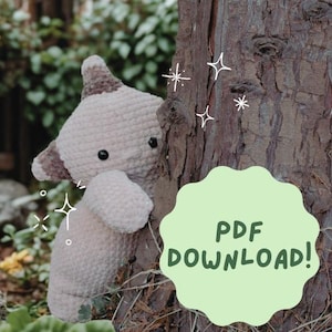 以下が含まれることがあります： ベージュと茶色の色合いの、黒いボタンの目と丸みを帯びた形の、かぎ針編みのぬいぐるみ。おもちゃは木の幹の隣に配置されています。テキスト「PDF DOWNLOAD！」が表示された緑色の吹き出し。