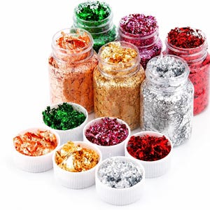 May include: A collection of colorful metallic flakes in various shades, including gold, silver, red, and green. The flakes are displayed in clear jars and small white bowls, creating a visually appealing arrangement. Ideal for craft projects.