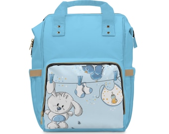 May include: A light blue diaper bag with a top handle and multiple pockets. The front pocket features a cartoon bunny, bird, baby socks, and a bib. The bag has a zipper closure and tan accents.
