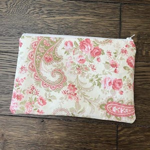 May include: A rectangular, cream-colored zippered pouch with a floral pattern. The pattern features pink roses, green leaves, and paisley designs. The zipper is white.