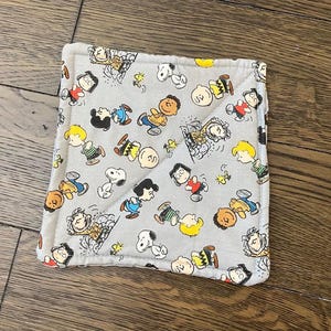 Puede incluir: Un salvamanteles cuadrado y acolchado de color gris claro con un patrón repetido de personajes de dibujos animados en varias poses. Los personajes son en tonos de negro, amarillo, rojo y marrón.