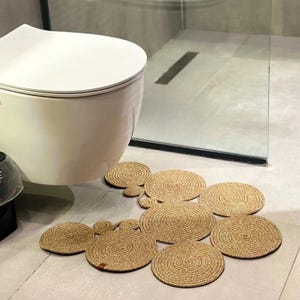 May include: A set of natural fiber bath mats in various sizes arranged in a cluster. The mats are circular with a woven texture and a light brown color. The mats are placed on a light gray tiled floor in a bathroom setting.