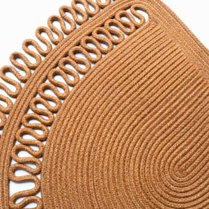 May include: Close-up of a brown woven rug with a decorative border. The rug has a circular design with a looped edge. The texture is visible, showing the individual fibers. The rug is made of natural fibers.