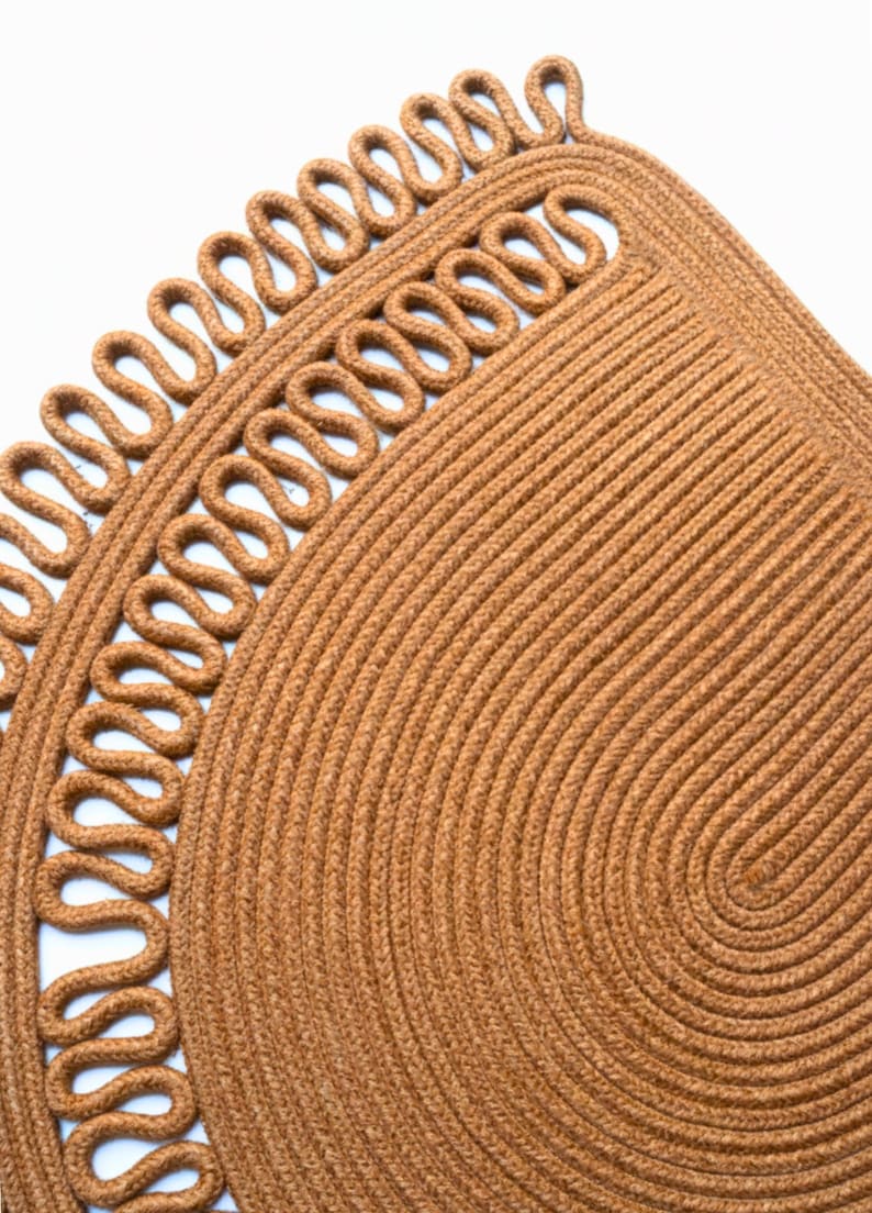 May include: Close-up of a brown woven rug with a decorative border. The rug has a circular design with a looped edge. The texture is visible, showing the individual fibers. The rug is made of natural fibers.