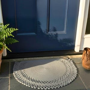 May include: A gray, semi-circular braided rug with a decorative edge sits on a slate tile surface in front of a navy blue door. A brown boot is visible to the right, and a potted fern is to the left.