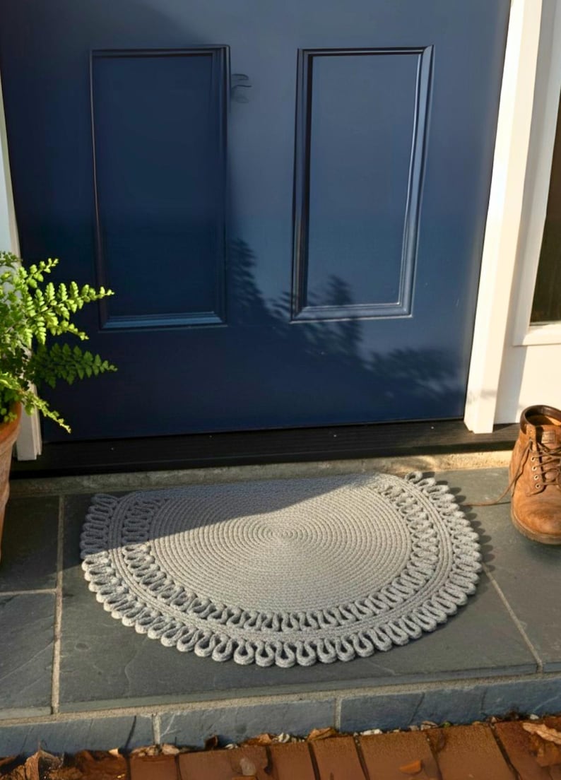 May include: A gray, semi-circular braided rug with a decorative edge sits on a slate tile surface in front of a navy blue door. A brown boot is visible to the right, and a potted fern is to the left.