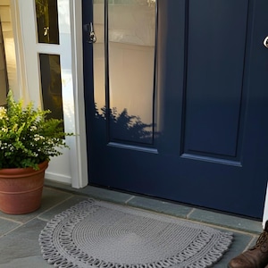 May include: A gray, semi-circular doormat with a decorative border sits in front of a navy blue door. The mat is made of a woven material. A potted plant and a pair of brown boots are also visible.