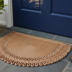 May include: A half-circle doormat made of natural jute fibers. The mat has a braided rope design with a scalloped edge. The doormat is placed in front of a blue door on a slate tile surface.