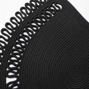 May include: Close-up of a black, circular woven placemat with a decorative looped edge. The placemat is made of thick, tightly woven material, creating a textured surface. The design features a spiral pattern in the center.