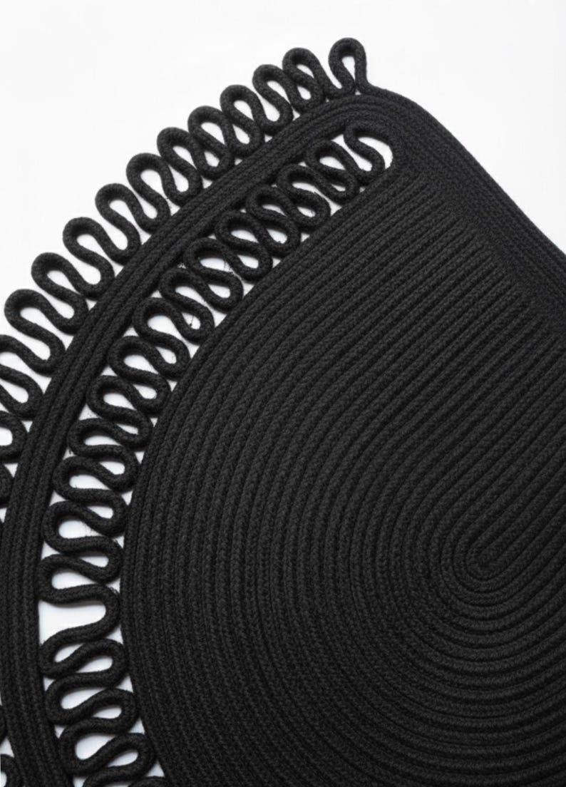 May include: Close-up of a black, circular woven placemat with a decorative looped edge. The placemat is made of thick, tightly woven material, creating a textured surface. The design features a spiral pattern in the center.