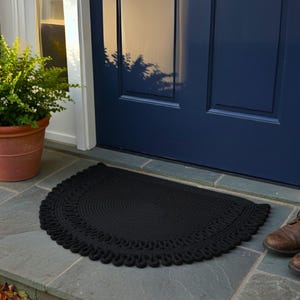May include: A black, semi-circular doormat with a decorative, looped edge sits on a stone surface in front of a blue door. A potted plant is to the left, and a pair of brown boots are to the right.