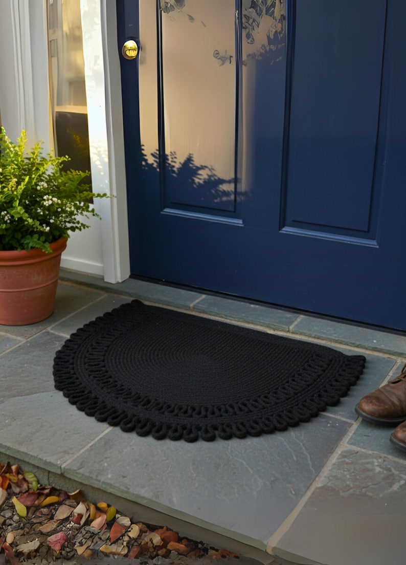 May include: A black, semi-circular doormat with a decorative, looped edge sits on a stone surface in front of a blue door. A potted plant is to the left, and a pair of brown boots are to the right.