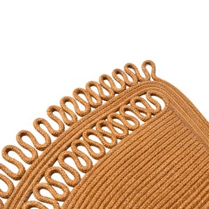 May include: Close-up of a tan-colored rug with a unique design. The rug features a repeating pattern of looped rope along the edge, creating a decorative border. The main body of the rug has a textured, woven appearance.