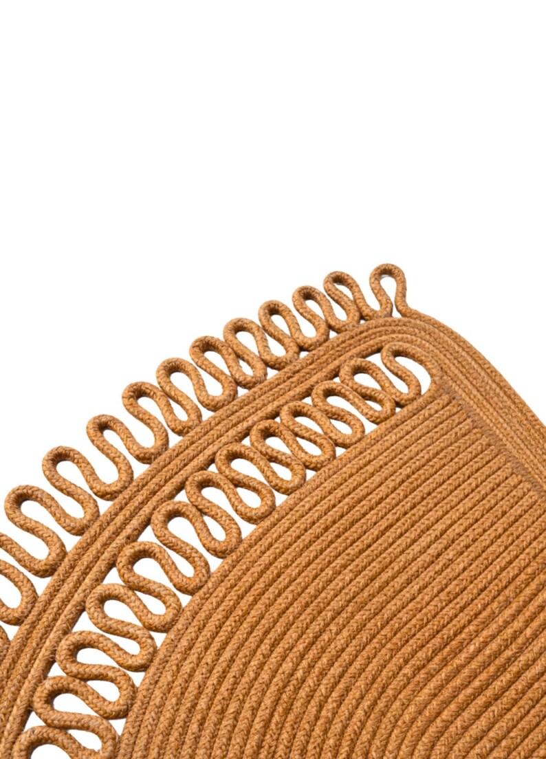 May include: Close-up of a tan-colored rug with a unique design. The rug features a repeating pattern of looped rope along the edge, creating a decorative border. The main body of the rug has a textured, woven appearance.