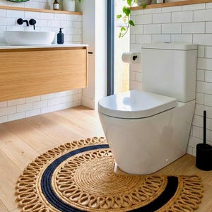 Könnte beinhalten: Ein runder Jute-Teppich mit einem marineblauen Streifen und einem dekorativen Schlaufenrand liegt auf einem hellen Holzboden in einem Badezimmer. Das Badezimmer verfügt über eine weiße Toilette, einen Holzwaschtisch und weiß geflieste Wände.