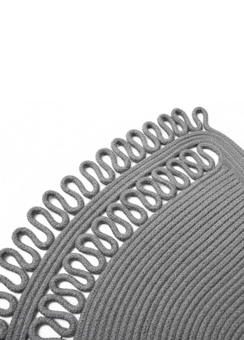 May include: Close-up of a gray, circular rug with a unique design. The rug features a looped border and a textured, braided pattern. The neutral color and simple design make it a versatile home decor item.