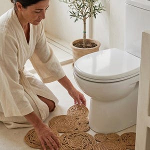 Może przedstawiać: Zestaw okrągłych, tkanych mat łazienkowych i podkładek z juty, ułożonych na jasnej podłodze łazienki. Maty mają spiralny wzór i są umieszczone w pobliżu białej toalety. Widoczne jest również małe drzewko oliwne w doniczce.