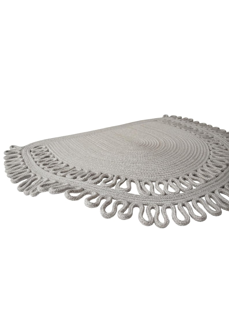 May include: A light gray, semi-circular rug with a looped edge design. The rug is made of a woven material and has a textured surface. The rug's diameter is approximately 30 inches.