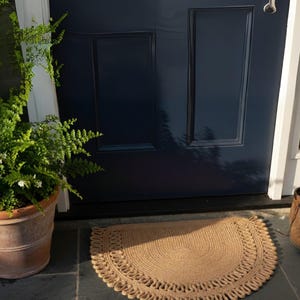 May include: A half-circle, tan jute doormat with a decorative border sits on a slate tile surface in front of a navy blue door. A potted fern is to the left of the mat.