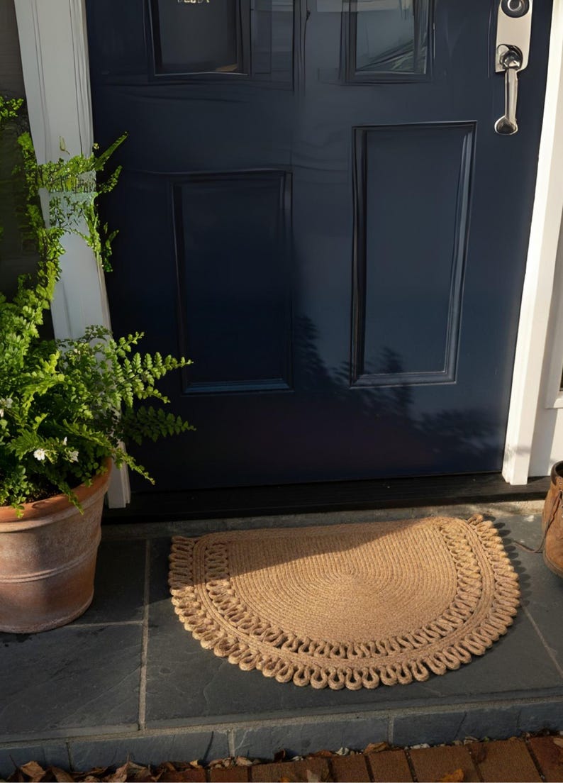 May include: A half-circle, tan jute doormat with a decorative border sits on a slate tile surface in front of a navy blue door. A potted fern is to the left of the mat.