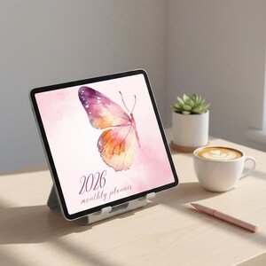 Op de afbeelding: Een digitale tablet met een aquarel vlinderontwerp in roze, paars en oranje. De tekst "2026 monthly planner" is zichtbaar. De tablet staat op een houten oppervlak, naast een kopje koffie en een pen.