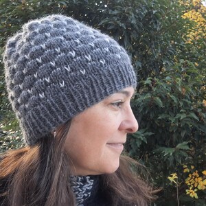 Puede incluir: Un gorro de punto gris con un patrón texturizado y detalles blancos. El gorro lo lleva una persona con cabello castaño, con un fondo de follaje verde y amarillo.