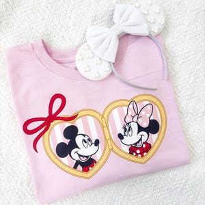 Op de afbeelding: Een lichtroze t-shirt met Mickey en Minnie Mouse in hartvormige medaillons. Een rode strik siert de shirt, en een witte strik haarband met parelaccenten ligt erbovenop. De shirt ligt op een wit, getextureerd oppervlak.