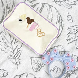 Puede incluir: Neceser crema con ribete morado, bordado con motivos de Mickey Mouse: helado, piruleta de chocolate, bebida rosa y pretzel. También hay una diadema azul claro con detalles de flores rosas.