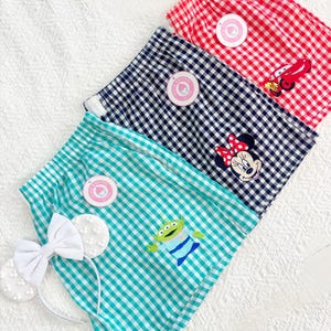 Op de afbeelding: Drie shorts met vichy-ruitjes in rood, marineblauw en talingblauw. De rode broek heeft een Lightning McQueen-afbeelding, de marineblauwe een Minnie Mouse-afbeelding en de talingblauwe een Alien-afbeelding. Een witte haarband met strik is inbegrepen.