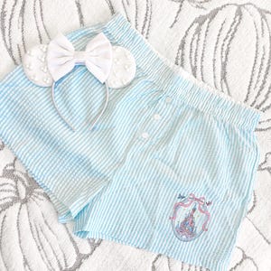 May include: Pale blue and white striped pajama shorts with a castle emblem and a white bow headband with mouse ears. The shorts have a button closure and are made of a soft, textured fabric.