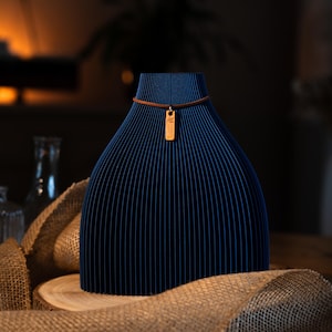 Può includere: Un oggetto decorativo blu scuro, a righe verticali, con una collana con cordino in pelle marrone e un ciondolo dorato. Il ciondolo è appeso davanti all'oggetto. L'oggetto è esposto su una superficie di legno con juta.