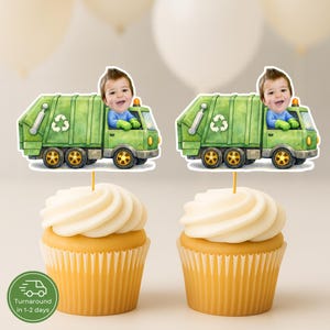 May include: Two cupcakes with white frosting, each topped with a decorative image of a green garbage truck with a child's face in the driver's seat. The cupcakes have yellow wrappers and are set against a blurred background.