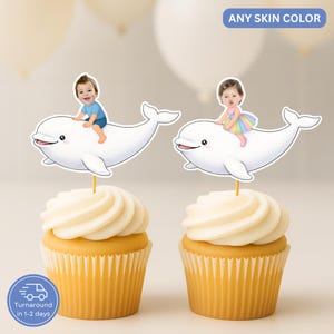 Pode incluir: Dois cupcakes com cobertura branca, cobertos com ilustrações personalizadas de crianças montando golfinhos brancos. O texto "ANY SKIN COLOR" está acima dos topos. Os cupcakes têm forros amarelos.