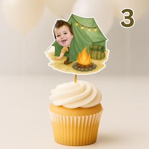 May include: A cupcake with white frosting topped with a camping-themed cake topper. The topper features a cartoon illustration of a child peeking out from behind a green tent with a campfire and the number 3.