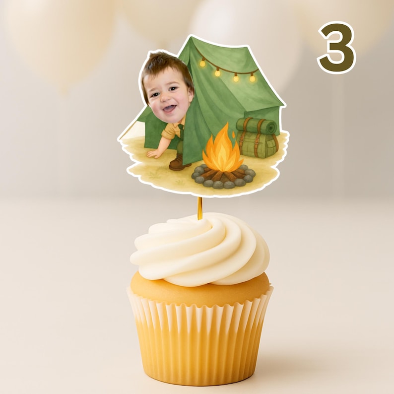 May include: A cupcake with white frosting topped with a camping-themed cake topper. The topper features a cartoon illustration of a child peeking out from behind a green tent with a campfire and the number 3.