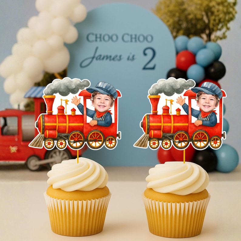 May include: Two cupcakes topped with personalized train-themed cake toppers. Each topper features a red steam locomotive illustration with a cutout of a child's face in the engineer's cabin, complete with a blue conductor hat and a puff of gray smoke.