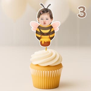 May include: A cupcake with white icing and a decorative topper. The topper shows a child's face in a bee costume holding a honey pot. The number "3" is visible in the background. The cupcake has a yellow paper case.