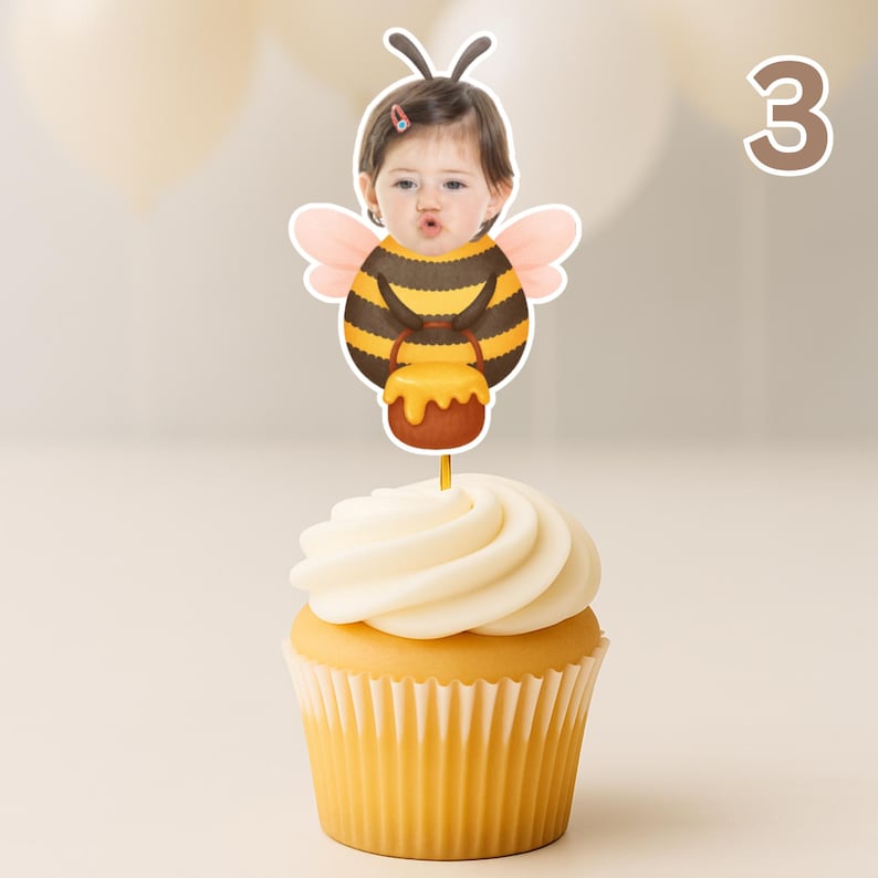 May include: A cupcake with white icing and a decorative topper. The topper shows a child's face in a bee costume holding a honey pot. The number "3" is visible in the background. The cupcake has a yellow paper case.