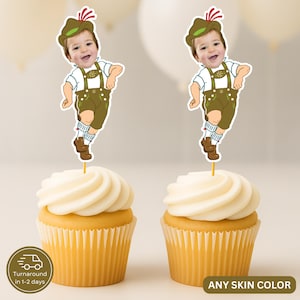May include: Two cupcakes topped with custom paper toppers featuring a photographic face of a child on a cartoon body wearing a green hat with a red feather, a white shirt, brown lederhosen with floral embroidery, white socks, and brown shoes.