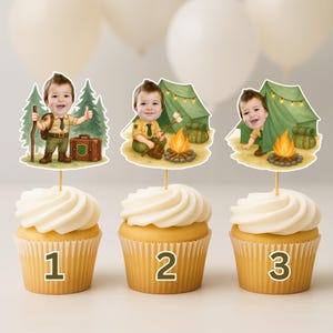 May include: Three cupcakes with white frosting, each topped with a cartoon image of a young boy in a scout uniform. The images depict camping scenes with a tent, campfire, and hiking gear. Numbers 1, 2, and 3 are printed on the cupcake wrappers.