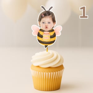 May include: A cupcake with white icing and a yellow paper case, decorated with a bee-themed cake topper. The topper shows a child in a bee costume holding a honey jar. The number "1" is visible in the background.