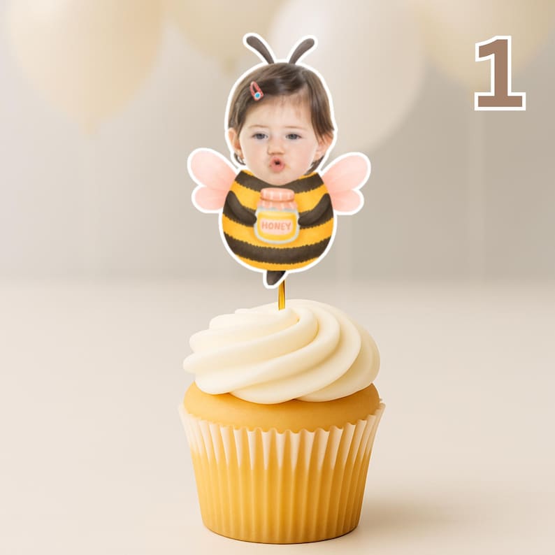 May include: A cupcake with white icing and a yellow paper case, decorated with a bee-themed cake topper. The topper shows a child in a bee costume holding a honey jar. The number "1" is visible in the background.