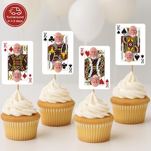 May include: Four cupcakes with white frosting, each topped with a playing card featuring a person's face as the King of various suits. The cards are on wooden skewers. The cupcakes sit on a white surface.