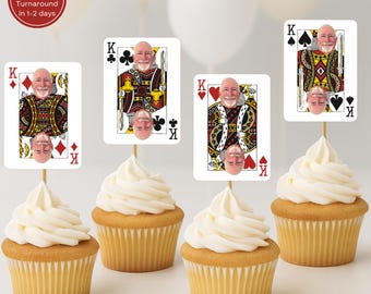 May include: Four cupcakes with white frosting, each topped with a playing card featuring a person's face as the King of various suits. The cards are on wooden skewers. The cupcakes sit on a white surface.